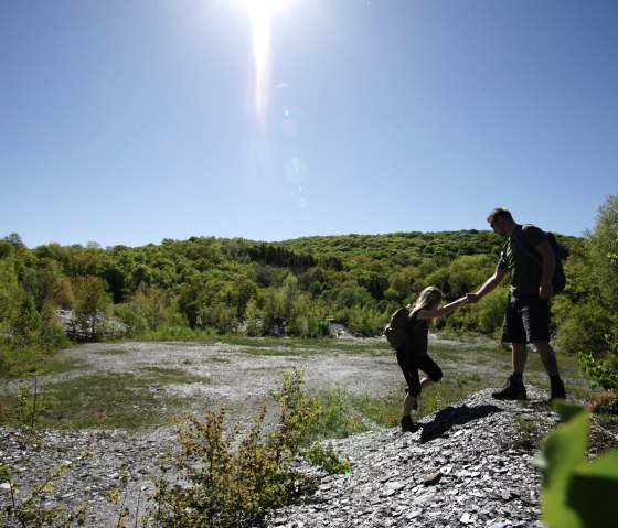 Twee wandelaars helpen elkaar een leistenen heuvel op in een groen landschap. De lucht is helder en zonnig., © Laura Rinneburger Twee wandelaars helpen elkaar een leistenen heuvel op in een groen landschap. De lucht is helder en zonnig., © Laura Rinneburger