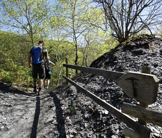 Twee wandelaars op een smal pad in een leistenen landschap, omringd door groene bomen en een blauwe lucht. Een houten bord wijst de weg., © Laura Rinneburger Twee wandelaars op een smal pad in een leistenen landschap, omringd door groene bomen en een blauwe lucht. Een houten bord wijst de weg., © Laura Rinneburger