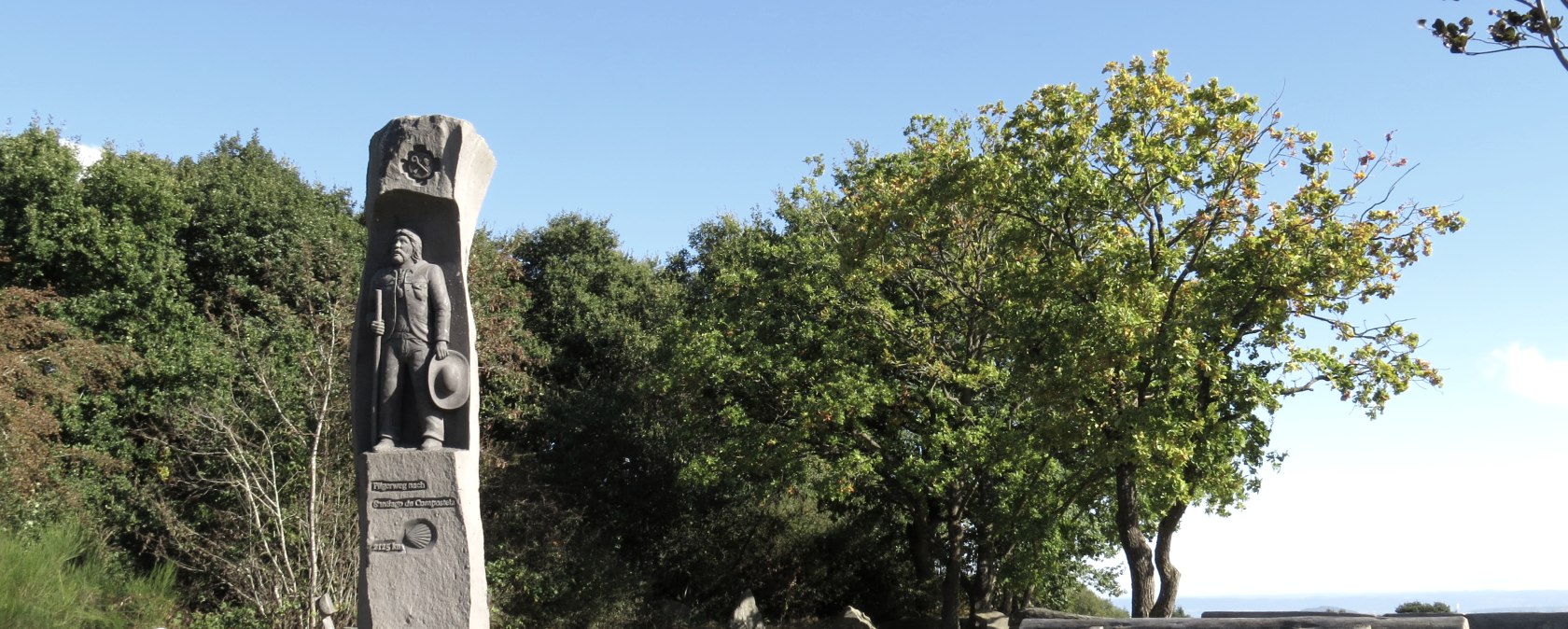 Die Jakobussäule auf dem Eifelcamino steht in einer grünen Landschaft. Daneben befindet sich eine rustikale Holzbank unter blauem Himmel. Die Jakobussäule auf dem Eifelcamino steht in einer grünen Landschaft. Daneben befindet sich eine rustikale Holzbank unter blauem Himmel.
