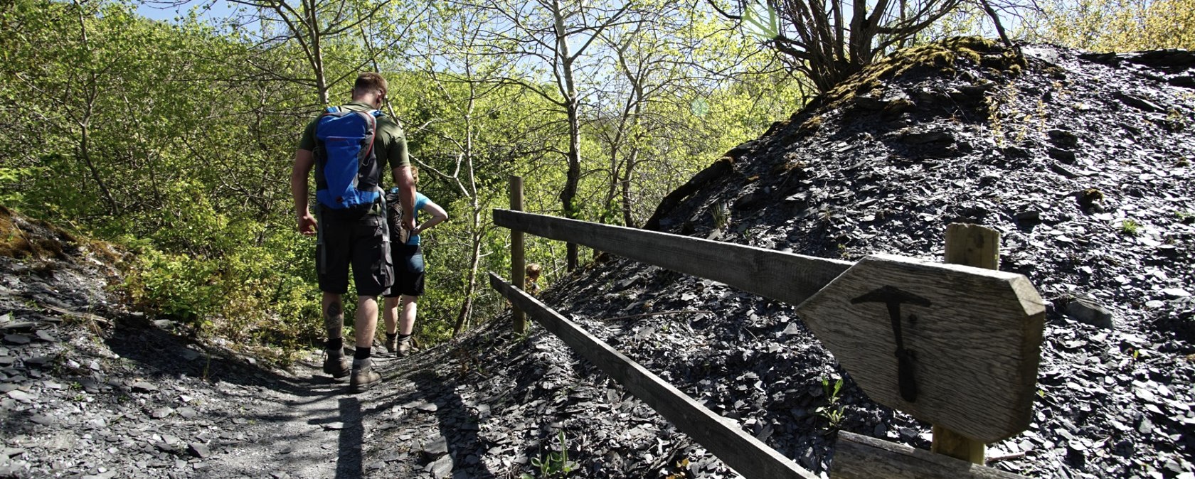 Twee wandelaars op een smal pad in een leistenen landschap, omringd door groene bomen en een blauwe lucht. Een houten bord wijst de weg., © Laura Rinneburger Twee wandelaars op een smal pad in een leistenen landschap, omringd door groene bomen en een blauwe lucht. Een houten bord wijst de weg., © Laura Rinneburger