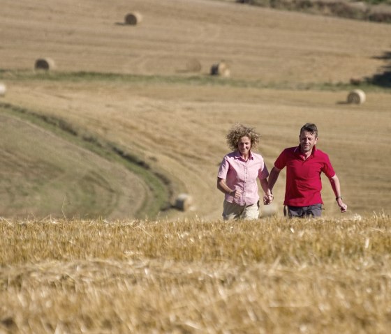 Ein Paar läuft Hand in Hand durch ein goldenes Feld. Im Hintergrund sind Heuballen auf den Feldern zu sehen., © Traumpfade/Kappest Ein Paar läuft Hand in Hand durch ein goldenes Feld. Im Hintergrund sind Heuballen auf den Feldern zu sehen., © Traumpfade/Kappest