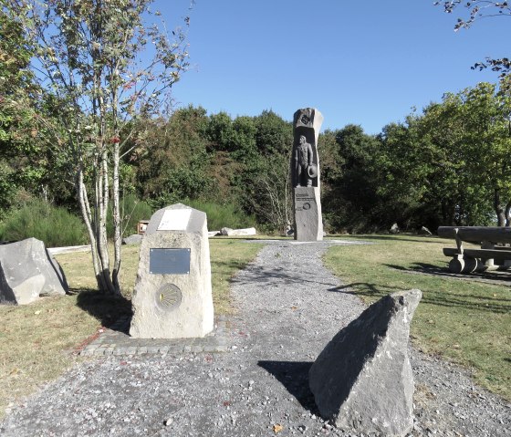 Ein Denkmal mit einer Statue und Informationstafeln auf einer grünen Wiesenfläche. Ein Picknicktisch steht rechts, umgeben von Bäumen unter klarem Himmel. Ein Denkmal mit einer Statue und Informationstafeln auf einer grünen Wiesenfläche. Ein Picknicktisch steht rechts, umgeben von Bäumen unter klarem Himmel.