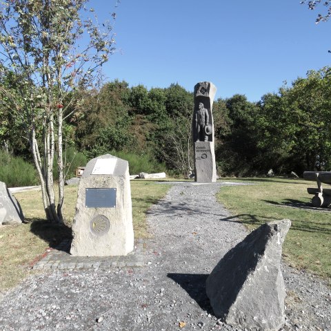 Ein Denkmal mit einer Statue und Informationstafeln auf einer grünen Wiesenfläche. Ein Picknicktisch steht rechts, umgeben von Bäumen unter klarem Himmel. Ein Denkmal mit einer Statue und Informationstafeln auf einer grünen Wiesenfläche. Ein Picknicktisch steht rechts, umgeben von Bäumen unter klarem Himmel.