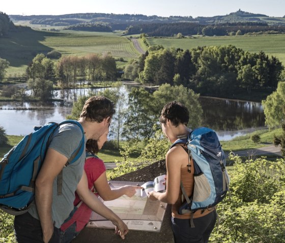 Drei Wanderer betrachten eine Infotafel vor dem Booser Doppelmaar. Im Hintergrund sind grüne Hügel und ein See zu sehen., © Kappest, REMET Drei Wanderer betrachten eine Infotafel vor dem Booser Doppelmaar. Im Hintergrund sind grüne Hügel und ein See zu sehen., © Kappest, REMET