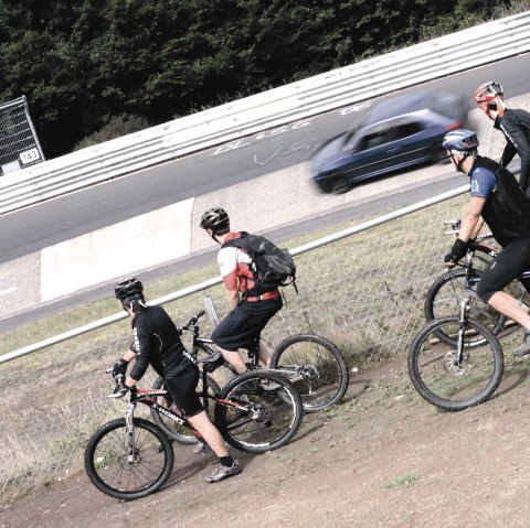 Vier Mountainbiker stehen an einem Zaun und beobachten ein Auto, das auf einer Rennstrecke vorbeifährt., © Markus Risse Vier Mountainbiker stehen an einem Zaun und beobachten ein Auto, das auf einer Rennstrecke vorbeifährt., © Markus Risse