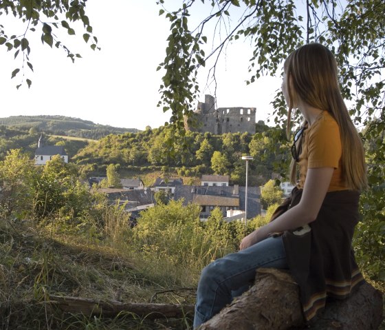 Eine Person sitzt auf einem Baumstamm und schaut auf eine Burgruine inmitten einer grünen Landschaft., © Traumpfade Eine Person sitzt auf einem Baumstamm und schaut auf eine Burgruine inmitten einer grünen Landschaft., © Traumpfade