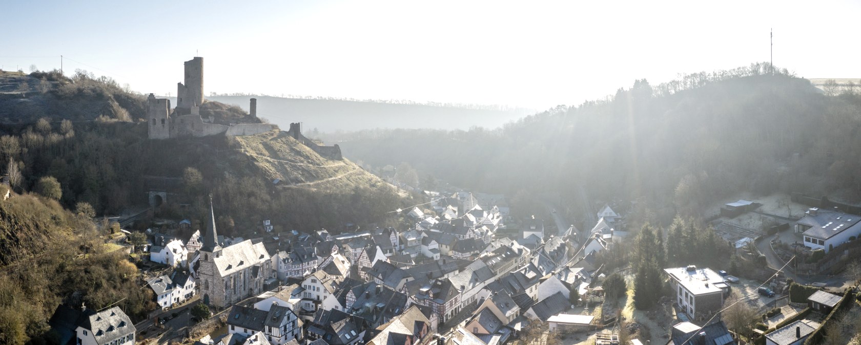 Luftaufnahme von Monreal, einem malerischen Dorf mit Fachwerkh&auml;usern und einer Burgruine auf einem H&uuml;gel im Hintergrund, umgeben von bewaldeten H&uuml;geln., &copy; Eifel Tourismus GmbH, D. Ketz
