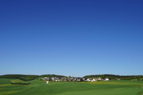 Eine grüne Landschaft mit sanften Hügeln und einem klaren blauen Himmel. Im Hintergrund sind kleine Häuser zu sehen.