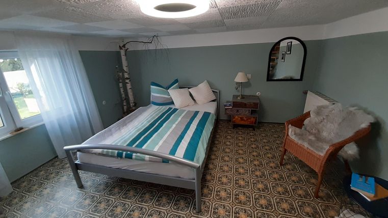 Ein einfaches Schlafzimmer mit einem Bett, das gestreifte Bettwäsche hat. Daneben stehen ein Nachttisch, eine Lampe und ein Sessel.