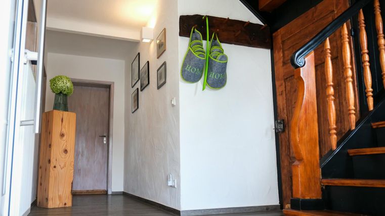 A bright hallway with a wooden staircase and modern decoration. Two green bags hang on the wall and a flower arrangement is in the corner.