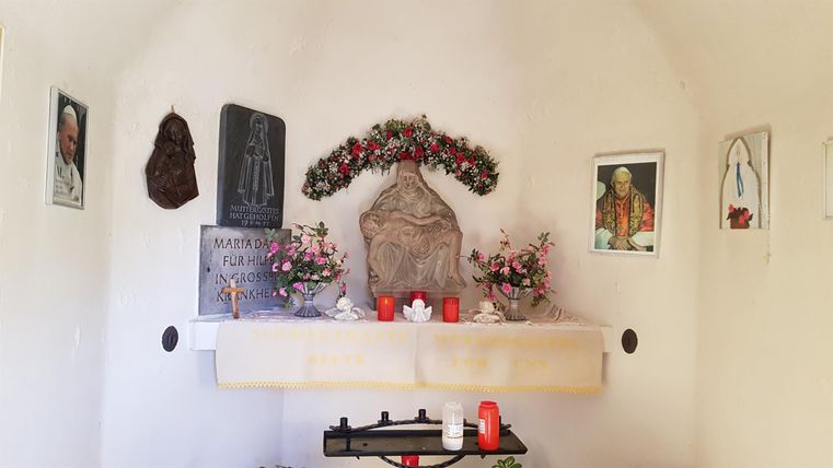 Ein kleiner, schlichter Raum mit einem Altar, der mit Blumen und Kerzen geschmückt ist. An den Wänden hängen verschiedene Bilder und Schrifttafeln.