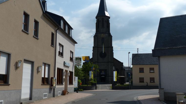 Eine ruhige Straße mit modernen Häusern und einer Kirche im Hintergrund. Der Himmel ist teilweise bewölkt.