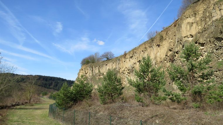 Eine steile Felswand mit grünen Bäumen am Fuß und einem klaren blauen Himmel. Der Weg führt durch eine ruhige Landschaft mit Wiesen und sanften Hügeln.