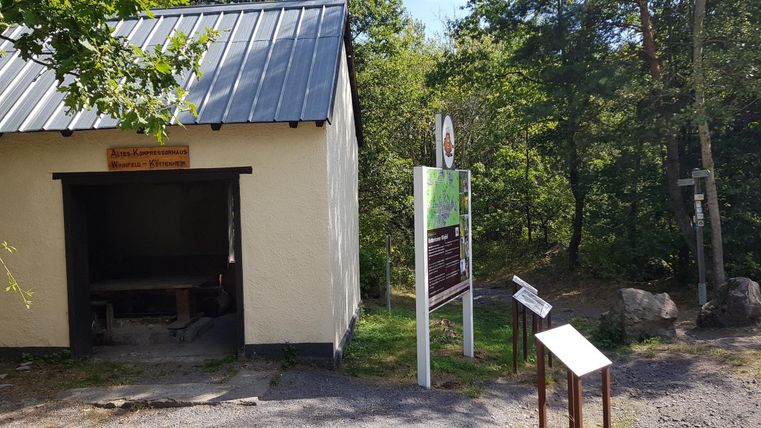 A small building with a metal roof and a wooden sign. Next to the building, there are information signs near a forest.