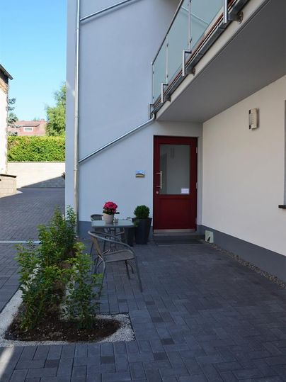 A modern entrance area with a red door and a small seating area. Surrounded by green plants and a paved path.