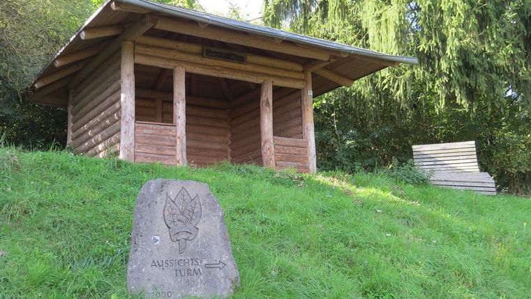 Ein kleiner Holzpavillon steht auf einer Wiese, umgeben von Bäumen. Ein Steinschild weist in Richtung eines Aussichtspunkts.