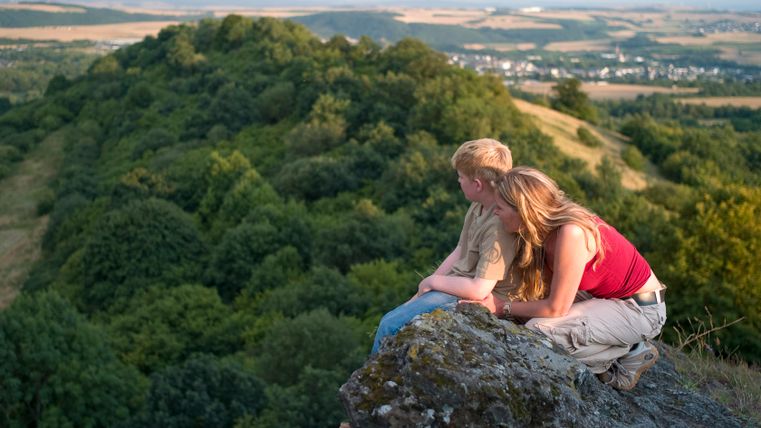 Two people sit on a rock with a view of a wooded landscape.