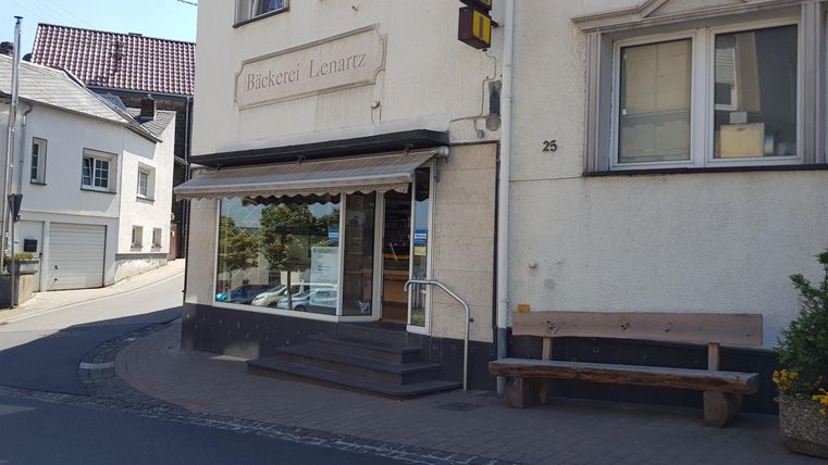 Ein kleines Geschäft an einer Straßenecke mit einer breiten Fensterfront. Draußen steht eine Holzbank und die Umgebung wirkt ruhig.