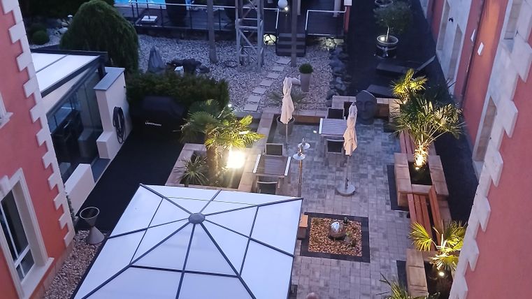 A beautiful courtyard with a pergola and comfortable seating. In the background, trees and a pool are visible.