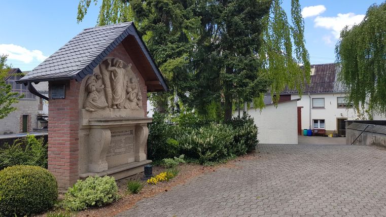 A beautifully designed sandstone fountain stands on a paved driveway. Surrounded by green plants and trees, it showcases a peaceful, rural setting.