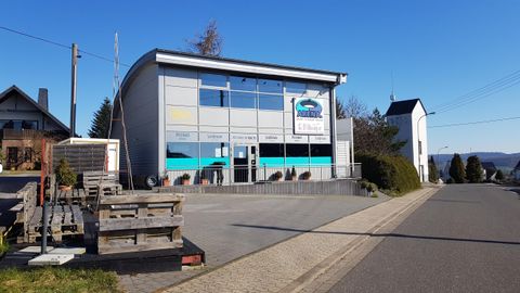 A modern building with large windows and a clear blue sky. In front of the building, some seating and a road are visible.