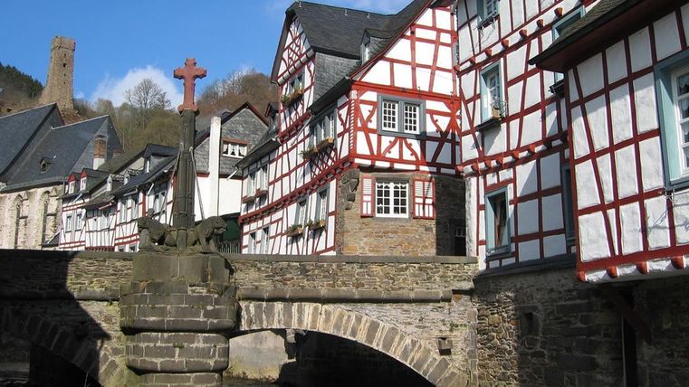 A picturesque town with half-timbered houses and a stone bridge over a small river. In the background, an old tower rises into the sky.