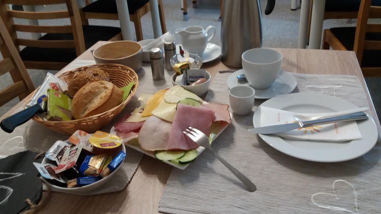 A laid breakfast table with rolls, cold cuts, and various jams. A coffee pot and dishes complete the picture.