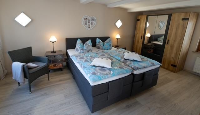 Ein gemütliches Schlafzimmer mit einem großen Doppelbett und bunten Bettdecken. Neben einem Sessel und einem Nachttisch sind Schränke im Hintergrund zu sehen.