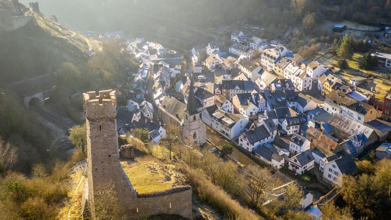 Eine historische Burg steht über einem malerischen Dorf. Die Häuser sind in sanften Farben gehalten, umgeben von hügeliger Landschaft.