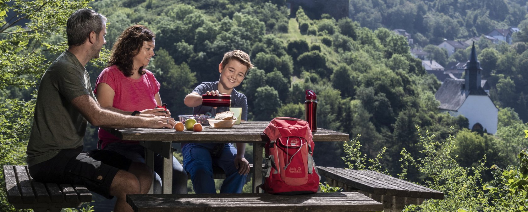 Familie beim Picknick an einem Tisch mit Blick auf eine Burgruine und eine Kirche in grüner Landschaft., © Kappest_Remet