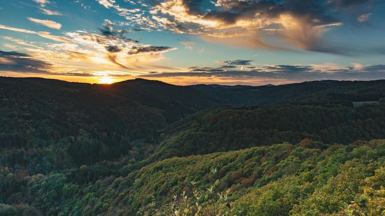 Eine wunderschöne Landschaft mit sanften Hügeln und bewaldeten Tälern. Der Sonnenuntergang beleuchtet den Himmel mit warmen Farben.