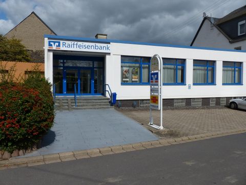 A modern bank building of the Raiffeisenbank with blue windows and an inviting entrance door. The sidewalk is paved and there is a green area in front of the building.