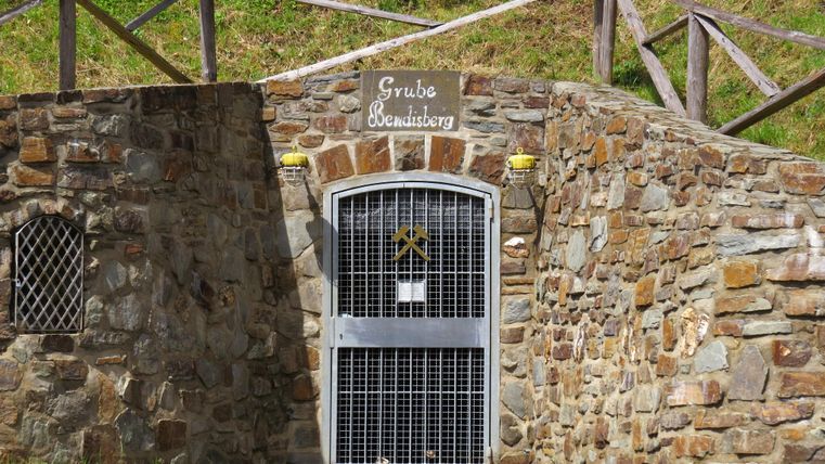 Ein Eingang zu einem alten Bergwerk aus Stein. Die Tür ist mit einem Gitter versehen und darüber steht der Name des Bergwerks.