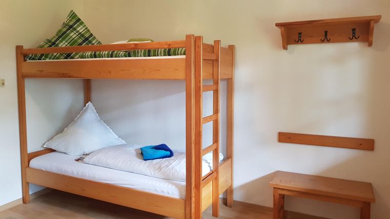 Ein einfaches Zimmer mit einem Etagenbett aus Holz. An der Wand hängen mehrere Haken und ein kleiner Tisch steht daneben.