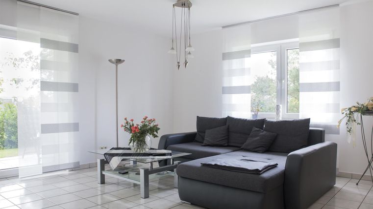 A modern living room with a gray sofa, a glass table, and large windows. The room has bright walls and minimal decoration.