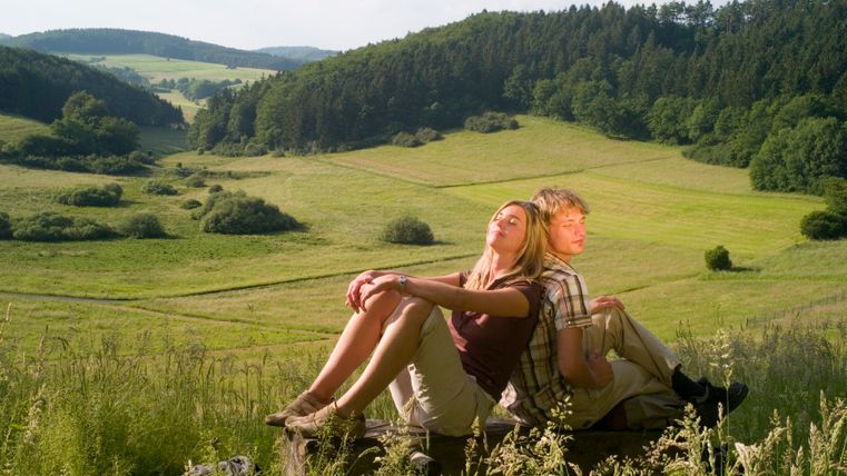 Zwei Personen sitzen auf einer Bank in einer grünen Landschaft mit Hügeln und Wäldern im Hintergrund.