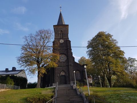Eine alte Kirche mit einem hohen Turm steht auf einem Hügel. Umgeben von Bäumen und einem blauen Himmel.