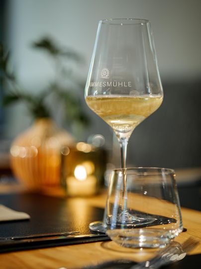 Ein elegantes Weinglas mit weißem Wein steht auf einem Tisch. Im Hintergrund sind verschwommene Kerzen und eine Pflanze zu sehen.