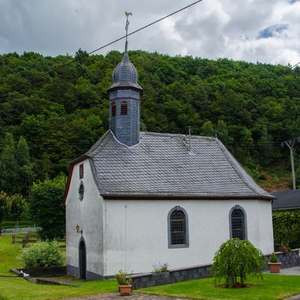 Kleine Kapelle mit grauem Schieferdach und Turm vor bewaldetem Hügel.
