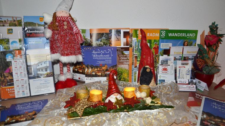 A festive decoration with Santa Clauses and candles is on a table. In the background, various brochures and flyers from a region can be seen.