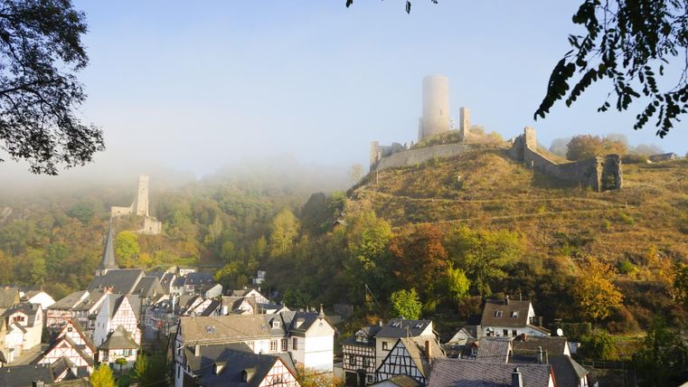 A picturesque landscape with an old castle on a hill and a misty surroundings. Below lies a charming village with traditional houses.