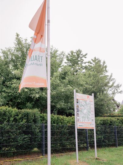 An orange flagpole stands next to an information sign. In the background, trees and a fence can be seen.