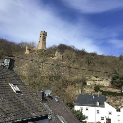 Eine alte Burgruine steht auf einem Hügel über einem Dorf. Der Himmel ist blau mit einigen Wolken.