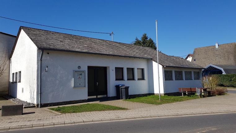 Ein einfaches, modernes Gebäude mit weißen Wänden und einem schrägen Dach. Es steht an einer Straße mit grünem Rasen und einem klaren, blauen Himmel im Hintergrund.