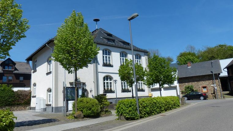 Ein modernes, zweistöckiges Haus mit einem hellen Anstrich und großen Fenstern. Umgeben von grünen Bäumen und einer ruhigen Straße.