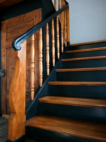 An interior staircase with wooden railings and black steps. The walls are plain, and in the background, a wooden door can be seen.