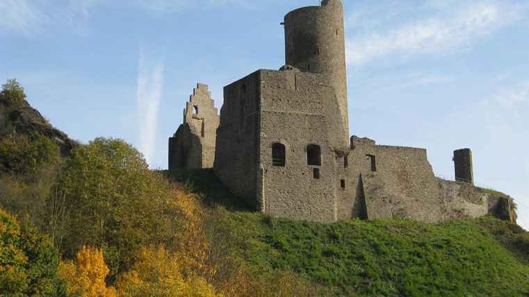 A medieval castle stands on a hill, surrounded by trees with colorful leaves. The sky is clear and blue.