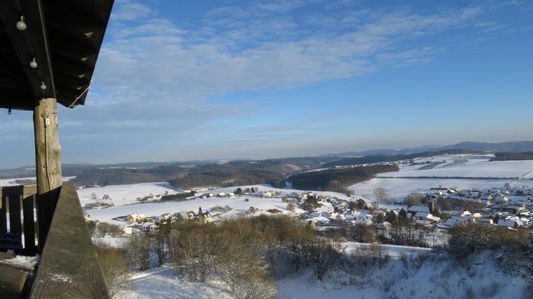 Eine winterliche Landschaft mit Schnee bedeckten Hügeln und einem klaren, blauen Himmel. Im Vordergrund ist ein Balkon mit Blick auf die schöne Aussicht.