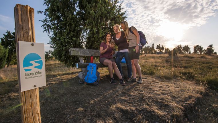 Three women sit on a bench in a heath landscape with a sign "traumpfädchen" in the foreground.