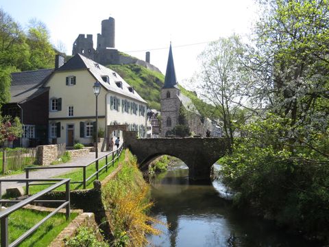A picturesque landscape with a small river and an old stone bridge. In the background, houses and a castle on a hill can be seen.